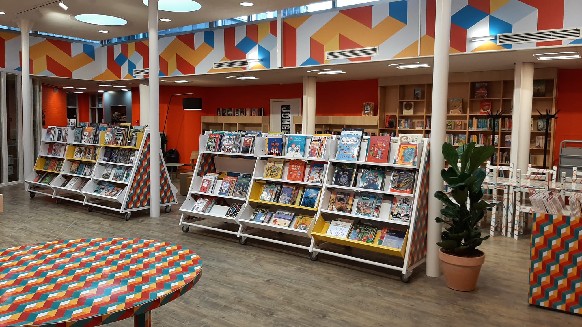 Een kleurrijk en modern interieurontwerp van een bibliotheekvestiging van de Bibliotheek Rotterdam. De ruimte heeft een lichte houten vloer, feloranje muren en een opvallende geometrische rand in primaire kleuren langs het plafond. In het midden bevindt zich een servicebalie met zelfbedieningsscanners en computers, voorzien van een speels patroon. Er staan kleurrijke zitbanken en boekenrekken met een open en uitnodigende opstelling. Silhouetten van mensen suggereren een levendige en toegankelijke bibliotheekomgeving.
